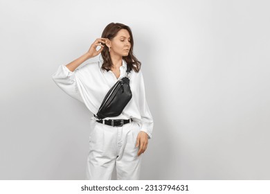 White Woman Wearing White Jeans, Shirt and Crossbody Black Leather Belt Bag over Grey Background