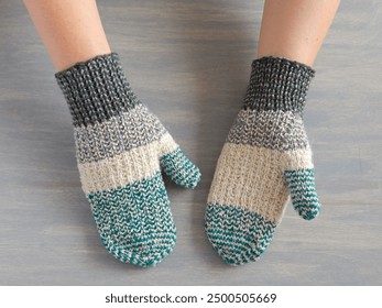 White woman's hands wearing handmade knitted multicolor scrap yarn mittens, grey background