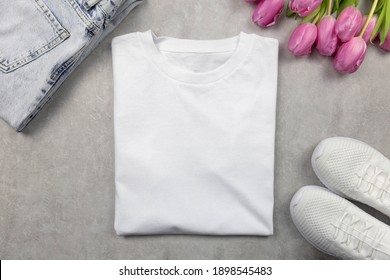 White Womens Cotton Tshirt Mockup With Pink Tulips, Jeans And Sneakers. Design T Shirt Template, Print Presentation Mock Up. Top View Flat Lay. Concrete Stone Background.