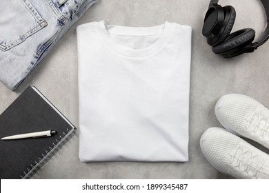 White Womens Cotton Tshirt Mockup With Jeans And Sneakers, Notebook And Black Headphones. Design T Shirt Template, Print Presentation Mock Up. Top View Flat Lay. Concrete Stone Background.