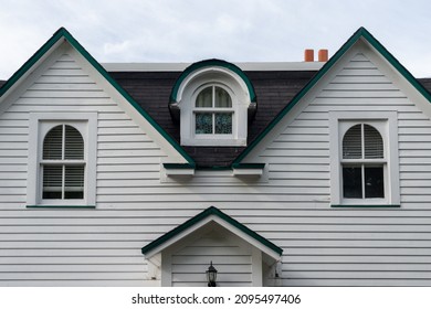 A white wood clapboard siding vintage house with multiple windows. The center window has a curved dormer over the closed glass window in the black shingled portion of the house. The sky is cloudy. 