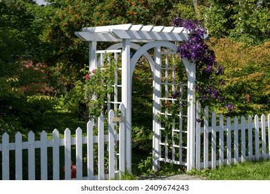 A white wooden archway and white wood picket fence surround a garden. There's a colorful purple vine hanging along the top of the fence and around the arch. The garden has trees and lush green grass. - Powered by Shutterstock