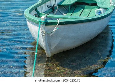 A white wooden boat, Rodney,  with green trim and interior. The rowboat is moored with a rope. The vessel is reflecting in the shallow clear water. There's a rope around the bow of the boat.