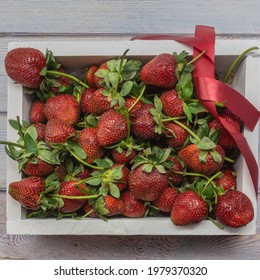 A white wooden box filled with ripe strawberries and tied with a red ribbon. Large berries with green leaves and cuttings. Close-up. Full screen. Top view