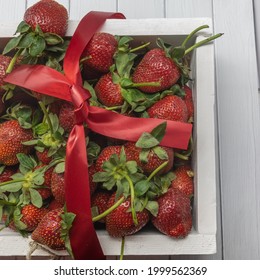 A white wooden box filled with ripe strawberries and tied with a red ribbon. Large berries with green leaves. Background - boards. Close-up. Top view