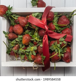 A white wooden box filled with strawberries and tied with a red ribbon. Ripe large berries with green leaves. Top view. Close-up