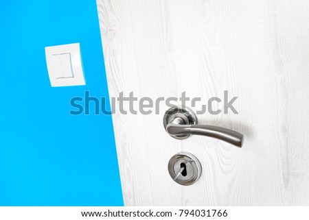 White wooden door with metal handle and keyhole with key in a bright mediterranean vivid light blue wall in a modern house with a small electricity light switch in a close up diagonal view