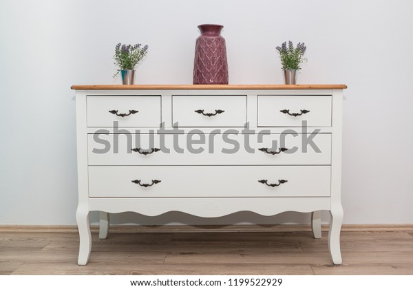 White wooden\
dresser with three vases and flowers on white wall background.\
Chest of drawers close\
up.