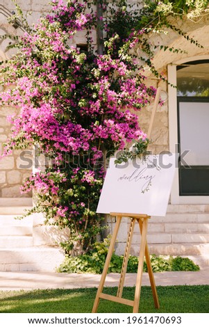 White wooden easel with an inscription on the background of a stone wall and a blooming rose bush. Title: Welcome to our wedding