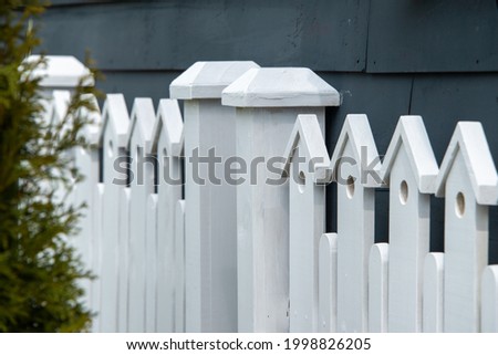 A white wooden fence with two gate posts in the center. The top of the fence has a small decorative birdhouse with a hole on the top of each paling.  There's a navy blue house in the background.