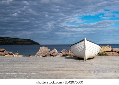 A white wooden fishing boat on the ocean shoreline under a blue sky with lots of clouds.  A land point is in the background meeting the horizon. The ocean is blue and calm with some gulls on rocks.