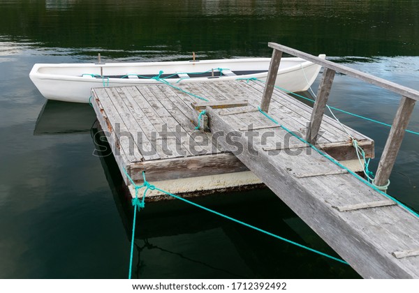 White wooden open boat tied to a small worn wooden\
floating dock and wharf. The water is blue with clouds reflecting\
in the water. The row bo is tied using a green rope. The water is\
calm and smooth. 