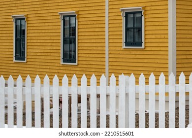 A White Wooden Picket Fence Is In The Foreground With A Bright Yellow Cape Cod Siding Covered Building In The Background. The House Has Multiple Vintage Windows In The Exterior Wall With Green Trim. 