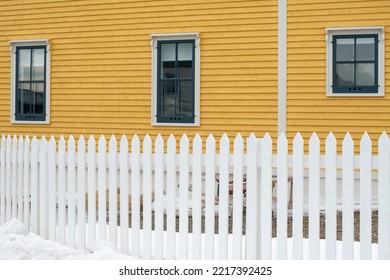 A White Wooden Picket Fence Is In The Foreground With A Bright Yellow Cape Cod Siding Covered Building In The Background. The House Has Multiple Vintage Windows On The Exterior Wall With Green Trim.