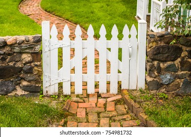 White wooden picket fence gate across a winding brick garden path in a historic neighborhood.