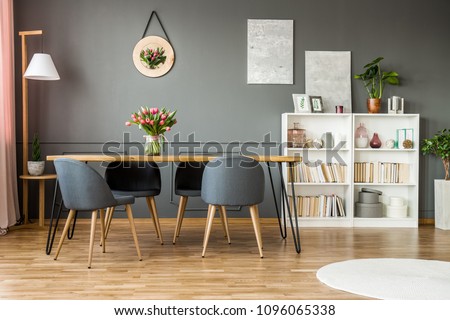 White wooden rack with books, decor and fresh plants standing in grey dining room interior with flowers on hairpin table