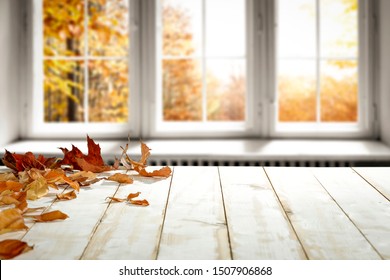 White Wooden Retro Table In Home Interior With Few Brown Leaves On Top. Blurred Background Of Window And Fall Landscape. Free Space For Your Decoration. 