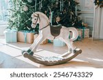 White wooden rocking horse in cozy festive living room on Christmas tree, surrounded by beautifully wrapped gift boxes. Winter holidays atmosphere. Wallpaper, background, backdrop