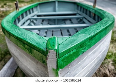A white wooden Rodney boat with bright green edging and a grey interior sits on the ground near the ocean. The bow of the dory is in the foreground. A traditional Newfoundland fishing boat newly built