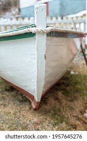 A white wooden Rodney boat with bright green edging and a grey interior sits on the ground near the ocean.The bow of the dory is in the foreground. A traditional Newfoundland fishing boat newly built.