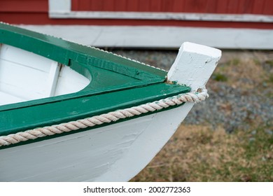 A white wooden Rodney boat with bright green edging and a grey interior sits on the ground near the ocean. The bow of the dory is in the foreground. A traditional Newfoundland fishing boat newly built