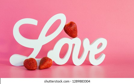 a white wooden sign saying LOVE with 1 strawberry leaning against the letters and two strawberries in front on a pink background