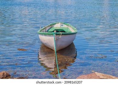A white wooden traditional dory or small fishing vessel with green trim sits on a smooth water surface. The dory has wooden oars and rope.  The smooth water is reflecting the image of the boat. 