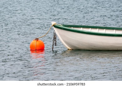 A white wooden traditional dory or small fishing vessel with green trim sits on a smooth water surface. The dory has wooden oars and rope.  The smooth water is reflecting the image of the boat. 