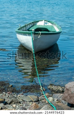 A white wooden traditional dory or small fishing vessel with green trim and interior is moored on a beach. The dory is tied to a large beach rock by a long green nylon fishing rope. 