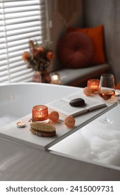 White wooden tray with glass of rose wine, book and burning candles on bathtub in bathroom