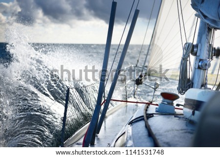 White yacht sailing in an open sea. View from the deck to the bow. Rough weather, storm, dramatic sky, dark clouds, waves, water splashes. Transportation, travel, sport, recreation, leisure activity