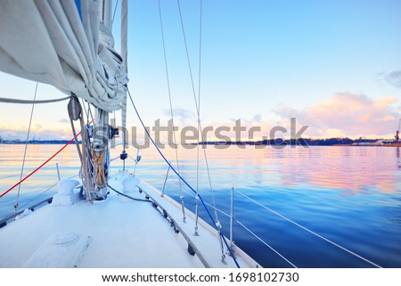 White yacht sailing in a still water at sunset. Frost and first snow on the deck, close-up view to the bow, mast, ropes and sails. Clear blue sky with colorful winter clouds. Norway