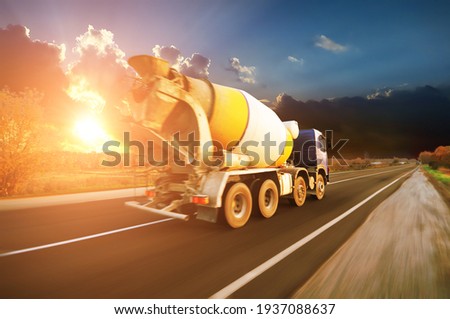 White and yellow concrete mixer truck on a countryside road in motion with trees against a blue sky with a sunset