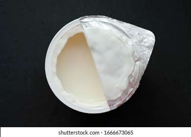 White yogurt in a plastic cup against a black background. Children's yogurt is half-open from above.
