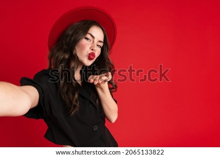 White young woman taking selfie photo while blowing air kiss isolated over red background
