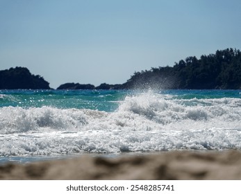 Whitecapped waves roll onto a light brown sandy beach under a clear blue sky. Dark green hills and rocky formations are visible in the distance. The sun is bright, indicating daytime. - Powered by Shutterstock