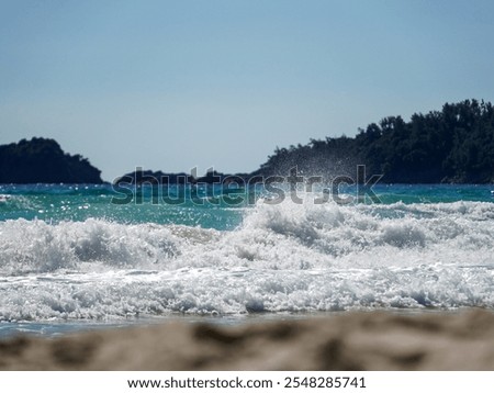 Whitecapped waves roll onto a light brown sandy beach under a clear blue sky. Dark green hills and rocky formations are visible in the distance. The sun is bright, indicating daytime.