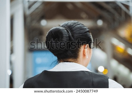 A whitecollar worker with black hair in a bun wearing a dress shirt is standing in front of an escalator, ready to travel to a formal event or job