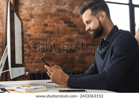 A whitecollar worker is seated at a desk using a cell phone, young handsome businessman scrolling news feed sittingin modern office space