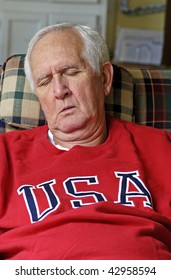 White-haired Senior man in bright red sweat shirt enjoys his retirement as he sleeps in his easy chair.