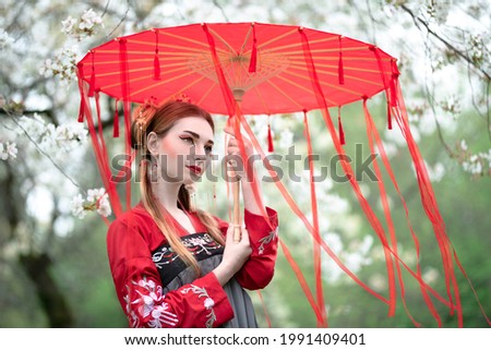 White-skinned girl in a beautiful hanbok under a red umbrella in a blooming spring garden