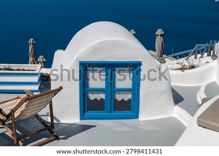 Whitewashed dome with bright blue window overlooking the Aegean Sea, Santorini, Greece, with terrace chairs and umbrellas in summer sunlight.