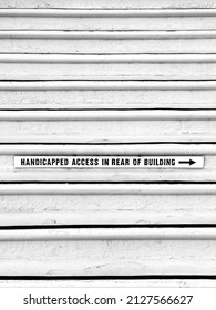 Whitewashed front stairway of commercial building with small sign on a riser partway up: "Handicapped access in rear of building." Black and white. Minimalist.
