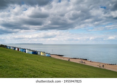 Whitstable, UK - April 4th 2021 Pretty Colourful Beach Huts Along A Seafront With Groynes. There Is Grass In The Foreground. People Are Walking And Cycling On The Promenade. The Sky Is Quite Cloudy.