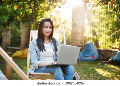 Who said you cannot work outdoors. Charming smiling girl in jeans, denim jacket sit comfortable picnic chair as studying in park, feet on grass, bean chair behind, girl connect wifi and use laptop.