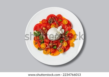 A whole ball of mozzarella lies on top of red and yellow tomato carpaccio with balsamic sauce and herbs in a ceramic, light plate on a gray background.