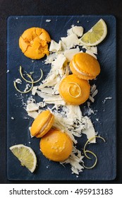 Whole And Broken Orange Lemon Homemade Macaroons With Chopped White Chocolate And Citrus Sugar And Zest On Dark Glass Board Over Black Textural Background. Overhead View