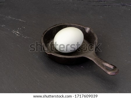 A whole chicken egg inside a tiny cast iron skillet on black background, viewed from above- inefficiency and incompetence concepts