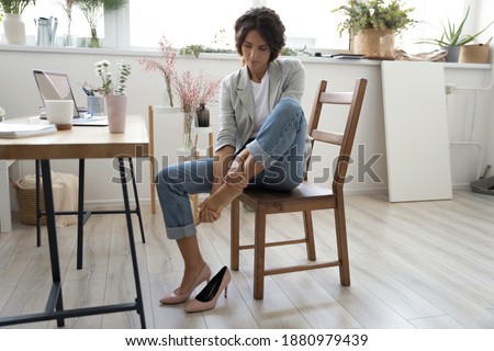 Whole day on stilettos. Tired young female florist decorator sitting on chair at work table taking off high heel shoe. Fatigued lady manager of craft store flower shop massaging bare foot feeling pain 商業照片 © 