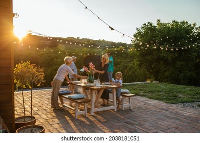 Whole family participates in the organization of dinner. Three female members setting dining table outdoors and male members barbecuing near them - Shutterstock ID 2038181051